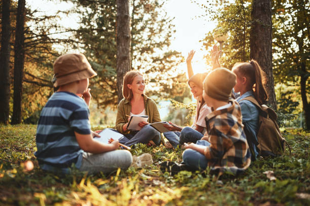 Des enfants et une femme assis sur l'herbe, dans une forêt, sous la lumière du soleil couchant.